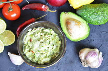 Bowl of freshly prepared guacamole. Ingredients on the background tomatoes, hot pepper, lime, avocado. Guacamole nachos and guacamole ingredients on dark wooden background.