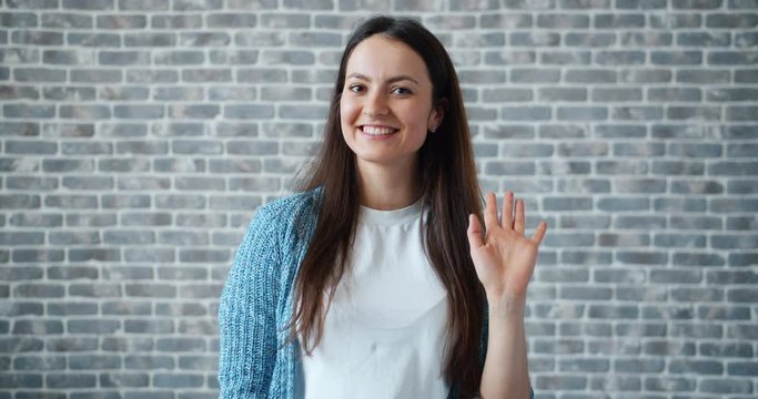 Portrait of friendy woman waving hand and looking at camera with happy face smiling greeting people on brick wall background. Welcome and hello concept.
