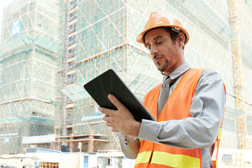 Serious young architect in hardhat and reflective vest standing outdoors and working online on digital tablet with high building in the background