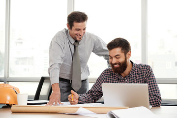 Bearded young businessman sitting at the table in front of laptop looking at blueprint and smiling while talking with his colleague at office