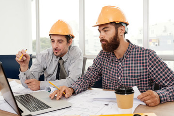 Two architects wearing hardhats sitting at the table using laptop computer and discussing working...