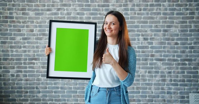 Portrait of girl pointing at green screen chroma key picture showing thumbs-up smiling standing on brick wall background holding frame. People and emotions concept.