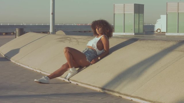 mixed race black young woman outdoors, summer sunset light, beach zone of Barcelona Panned video model sit on concrete skaters structure