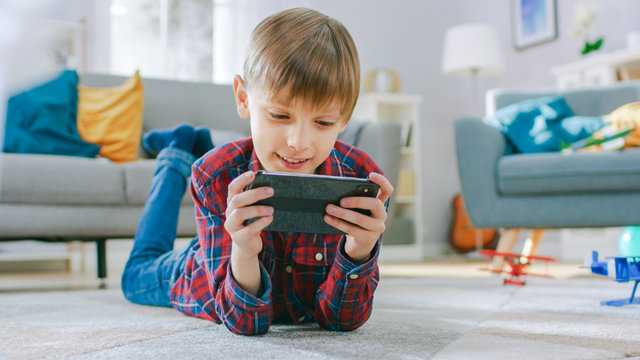 Smart Little Boy Laying On A Carpet Plays In Video Game On His Smartphone, Holds Mobile Phone In Horizontal Landscape Mode. Child Has Fun Playing Videogame In Sunny Living Room.