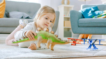 Cute Little Girl Blond Laying on a Carpet at Home, Plays with Toy Dinosaurs and Airplanes. Happy...