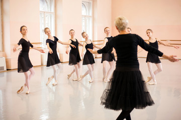 Senior ballet instructor demonstrating moves in front of a group of teenage girls. Practicing classical ballet in a large dancing studio.