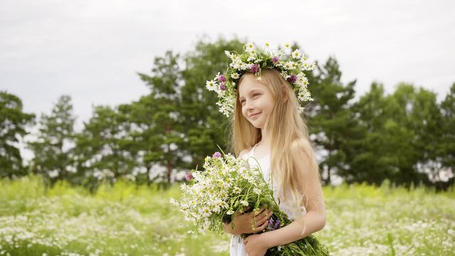 Portrait of beautiful girl is standing on flower glade with closed eyes. Cute teen girl in white dress with wreath on head and bouquet of daisies at flower meadow. Windy summer day.