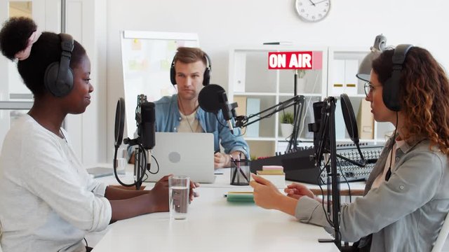 Zoom out of three diverse radio hosts sitting at microphones at table and communicating on air
