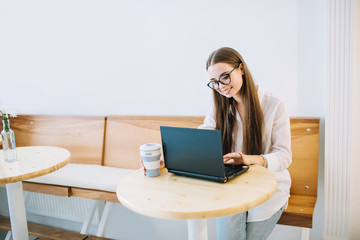 Young smiling woman working on laptop at a cafe, while drinking coffee. Young attractive girl sitting in a coffee shop with black laptop.