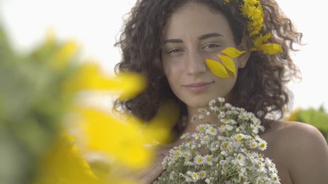 Portrait of a confident beautiful curly girl looking at the camera smiling standing in the sunflower field with bouquet of wildflowers. Bright yellow color. Freedom concept. Happy woman. Slow motion.