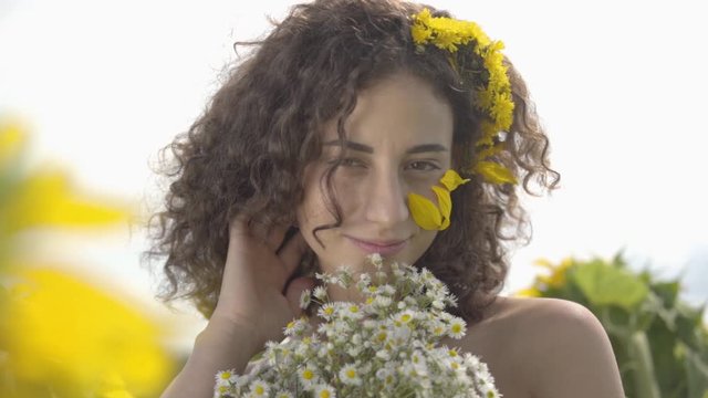 Portrait of a cute curly girl looking at the camera smiling standing in the sunflower field with bouquet of wildflowers. Bright yellow color. Freedom concept. Happy woman outdoors. Slow motion.