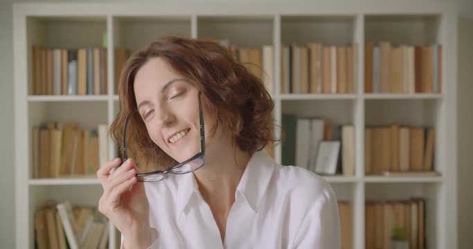 Closeup portrait of adult redhead attractive caucasian businesswoman holding her glasses looking at camera indoors with bookshelves on the background