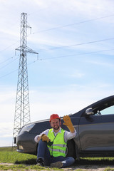 An electrician in the fields near the power transmission line. The electrician manages the process of erecting power lines. The mechanic in a helmet and a reflective form special gloves at work.