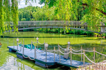 Obraz premium View of wooden pier, decorated with lights and rope, in calm green lake water with bridge on background