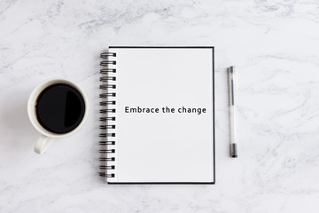 Embrace the change writing on notebook. Notebook on desk with coffee cup and a pen.