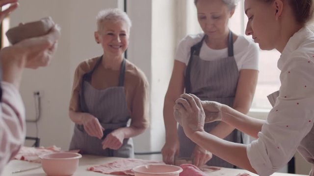 Tilt up of young female tutor standing at wooden table in ceramic workshop and showing middle-aged women how to shape clay then giving sample to one of women