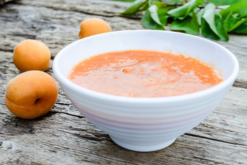 Apricot jam in a white bowl next to ripe apricot and green leaves on a wooden surface.