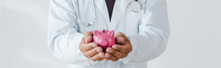 panoramic shot of doctor holding pink piggy bank in hands on white