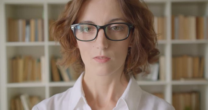Closeup portrait of adult redhead attractive caucasian businesswoman in glasses looking at camera indoors in library bookshelves on background