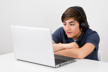 Smiling caucasian teenage kidy with headphones sitting at the table and having fun while watching a video at his laptop