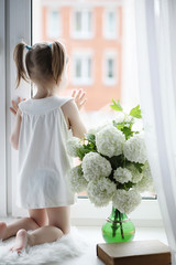 A little girl is sitting on the windowsill. A bouquet of flowers in a vase by the window and a girl sniffing flowers. A little princess in a white dress with a bouquet of white flowers by the window.