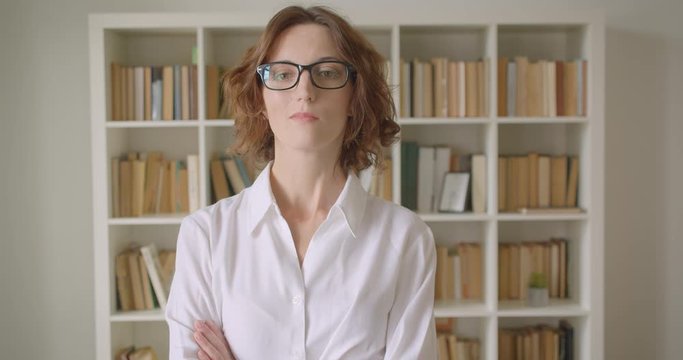 Closeup portrait of adult redhead caucasian businesswoman in glasses looking at camera with her arms crossed over chest indoors with bookshelves on the background