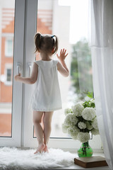 A little girl is sitting on the windowsill. A bouquet of flowers in a vase by the window and a girl sniffing flowers. A little princess in a white dress with a bouquet of white flowers by the window.