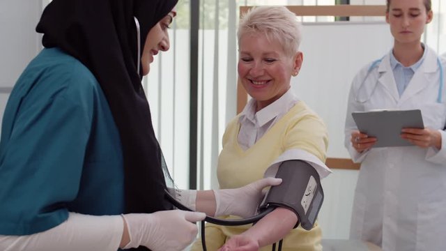 Tilt up of female Muslim doctor wearing veil measuring blood pressure of middle-aged blonde Caucasian woman sitting on daybed and then telling results to her female colleague making notes in card