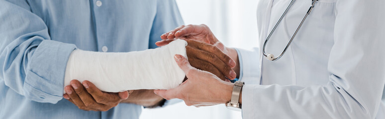 panoramic shot of doctor touching broken arm of man in clinic