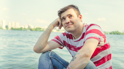 Man by the river.Portrait of a male in a t-shirt sitting on a column near the river on a Sunny day