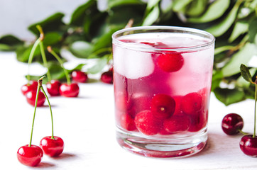 A glass of cherry drink with ice in a glass on a white wooden background. Fresh summer cocktail with cherries and ice cubes.