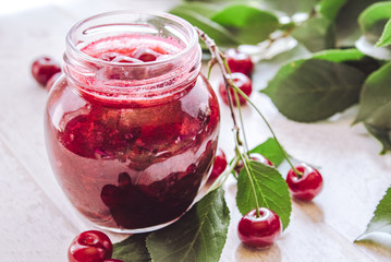 Bank with homemade cherry jam on a wooden background near the berries and leaves.