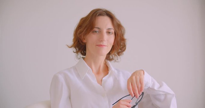 Closeup portrait of redhead caucasian businesswoman looking at camera smiling happily holding glasses with thoughtfulness sitting in armchair white office