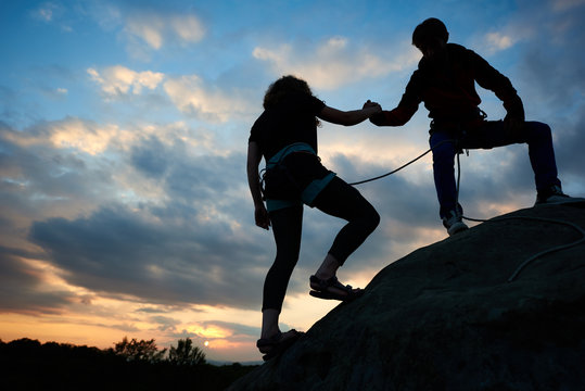 Young male and female climbers helping each other to conquer the summit in teamwork on fantastic mountain landscape at sunset. Couple silhouette on the top of rock with wonderful heaven on background.