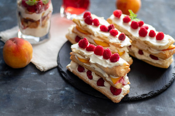 Dessert Milfey. Strips of puff pastry with curd cream raspberry berries and peach slices. In the background, the same, only in a glass. On a dark background. 