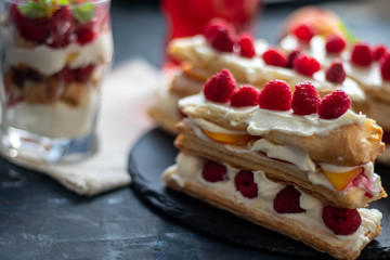 Dessert Milfey. Strips of puff pastry with curd cream raspberry berries and peach slices. In the background, the same, only in a glass. On a dark background. 