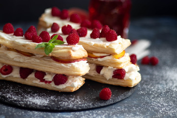 Dessert Milfey. Strips of puff pastry with curd cream raspberry berries and peach slices. In the background, the same, only in a glass. On a dark background. 