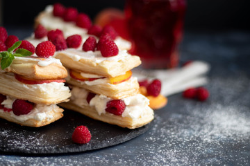Dessert Milfey. Strips of puff pastry with curd cream raspberry berries and peach slices. In the background, the same, only in a glass. On a dark background. 