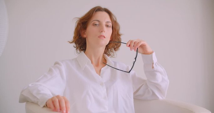 Closeup portrait of redhead caucasian businesswoman looking at camera holding glasses with thoughtfulness sitting in armchair white office