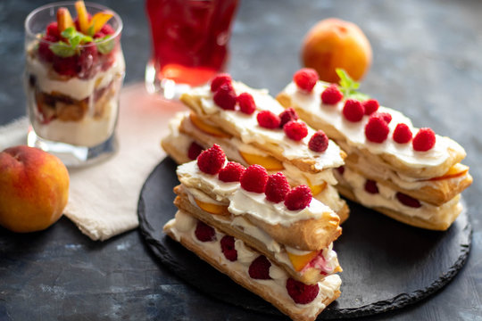 Dessert Milfey. Strips of puff pastry with curd cream raspberry berries and peach slices. In the background, the same, only in a glass. On a dark background. 