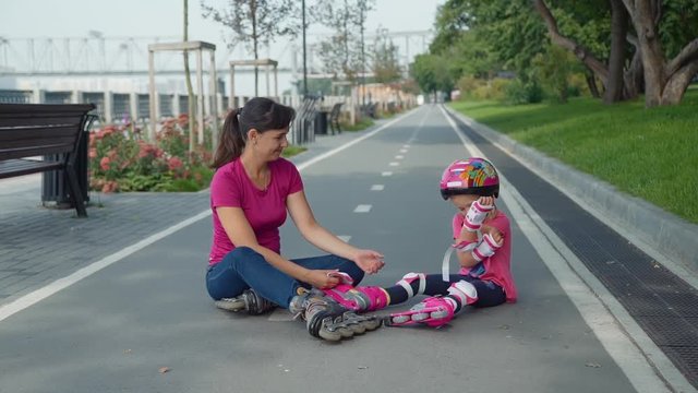 Mother and Daughter Sitting on the Bicycle Lane after Rollerblading in a Park. Little Girl in the Colorful Sport Helment Taking Off her Protective Gear with Help of her Mother. Slow Motion.