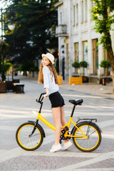 Fototapeta na wymiar Girl teenager in a hat with a yellow bike walks through the summer city.