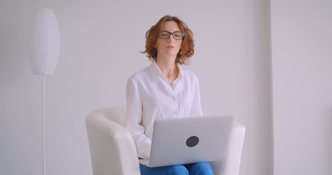 Closeup portrait of adult redhead caucasian businesswoman in glasses using the laptop looking at camera sitting in the armchair in the white office