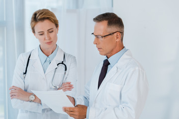 man in glasses and white coat looking at blank paper near coworker with crossed arms