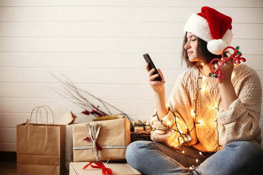 Stylish happy girl in santa hat looking at phone screen in festive christmas lights on background o presents and gifts in modern room. Young hipster woman in cozy sweater browsing online