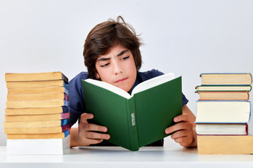 Teenage kid with serious concentrated expression sitting at the desk between pile of books and reading a book on capitalism