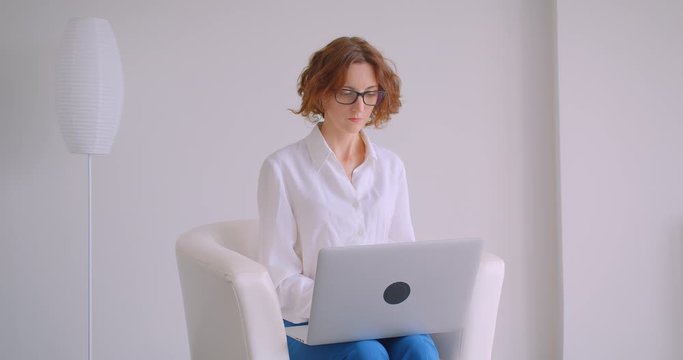 Closeup portrait of adult redhead caucasian businesswoman in glasses using the laptop looking at camera smiling happily sitting in the armchair in the white office
