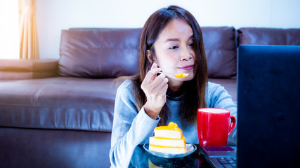 Beautiful Asian women sitting in the living room. She eating orange cake with coffee  and sit and check the work on the computer. Concept Working outside the office via the internet.
