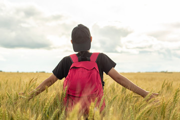 A teenager with a backpack stands in a wheat field (Triticum) and touches the ear with his hands, against the sky and clouds. Summer in the village. Holidays. The concept of freedom and relaxation.
