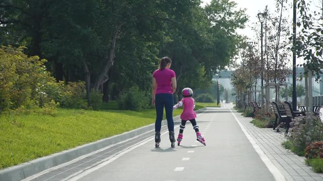 Young Mother Roller Skating with her Daughter Outdoors. Young Woman Teaching Little Girl to Rollerblading in Sunny Day. Summer Family Activities Concept